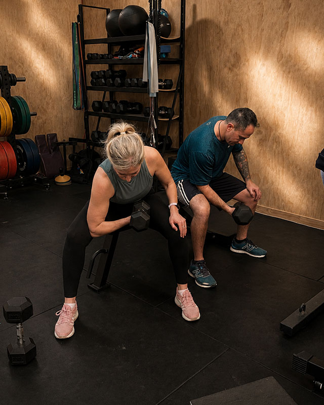 Two athletes training together with one coach in an industrial gym, partner-assisted kettlebell exercise, encouraging atmosphere, natural light, editorial fitness photography, muted tones with warm gold accent