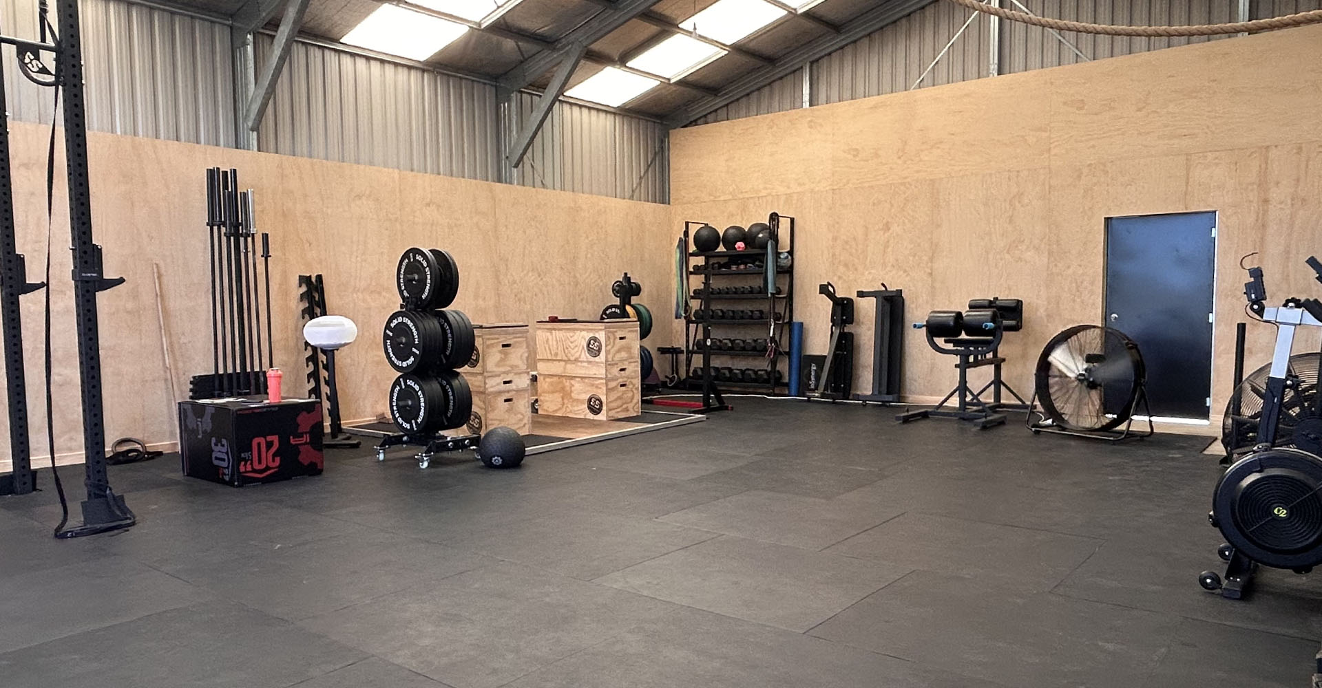 Coach and prospective client shaking hands and talking beside a lifting platform in a converted shed gym, warm evening light through a roller door, genuine and welcoming atmosphere, editorial fitness photography