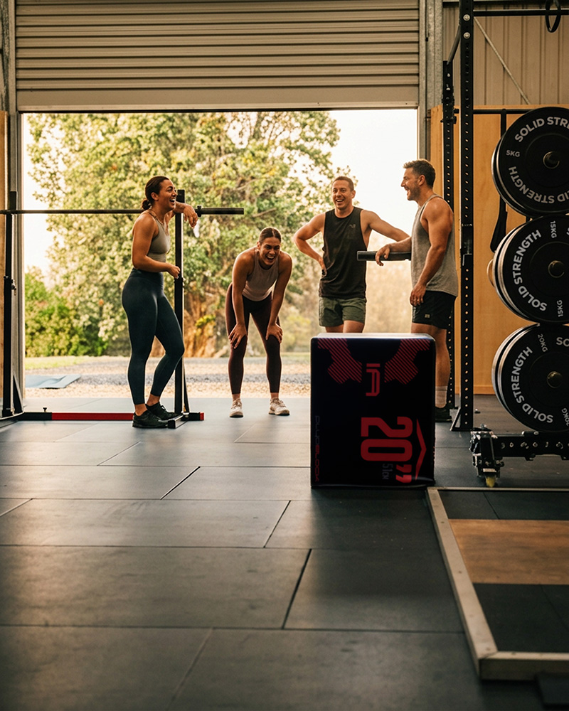 Vertical shot of two or three athletes standing close together in a tight huddle, laughing and catching their breath between sets in a converted shed gym, genuine candid moment, kettlebell visible in the foreground, warm late-afternoon light through an open roller door, editorial lifestyle photography, portrait orientation