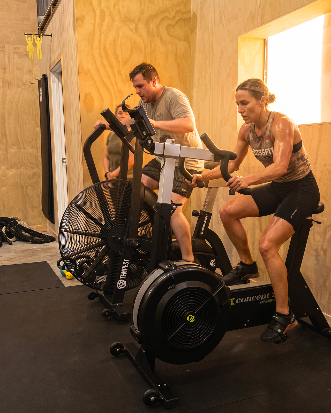Vertical, tightly framed shot of two group class participants of visibly different fitness levels performing a scaled version of the same exercise close together — one with a lighter kettlebell in the foreground, one with a heavier kettlebell just behind, coach's hand or arm visible guiding from the side, inclusive and encouraging atmosphere, editorial fitness photography, natural light, portrait orientation