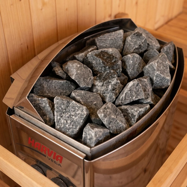 Close-up detail shot of sauna stones on a heater with a wooden ladle and bucket beside it, warm low light, steam rising gently, intimate still-life wellness photography